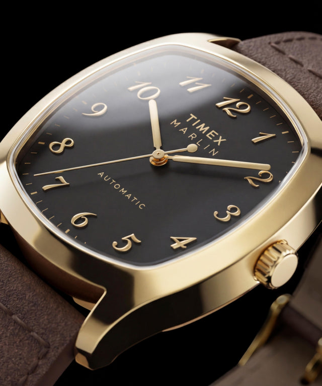 Gros plan de la Timex Marlin Chatham Automatic dorée, montrant son cadran noir, ses chiffres arabes appliqués dorés, ses aiguilles dorées et son bracelet en cuir marron
