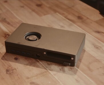Une console de jeux noire compacte, dotée d'un lecteur de disque intégré, de ports USB et d'un ventilateur de refroidissement situé sur le dessus, posée sur une table en bois.