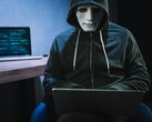 Un personnage masqué et encapuchonné travaille sur un ordinateur portable dans une pièce sombre.