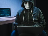 Un personnage masqué et encapuchonné travaille sur un ordinateur portable dans une pièce sombre.