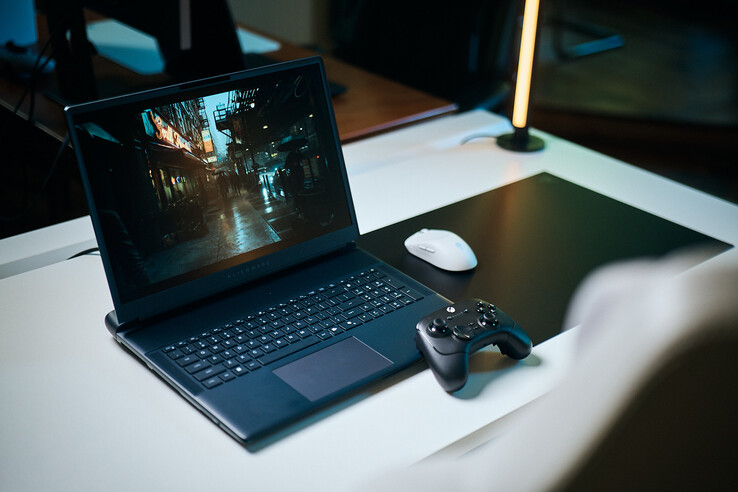 Un ordinateur portable de jeu Alienware ouvert, posé sur un bureau de studio blanc, affichant une scène de rue sombre et réfléchissante avec des détails nets obtenus par le « path tracing », accompagné d’une manette noire et d’une souris sans fil blanche.