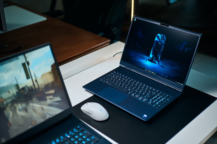 Un ordinateur portable de jeu de milieu de gamme bleu foncé (Alienware Aurora 16X) posé en biais sur un tapis de bureau arborant un fond d'écran de science-fiction bleu lumineux, placé à côté d'une souris sans fil blanche, avec un autre ordinateur portable flou au premier plan.