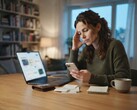 Une femme assise à un bureau regarde son smartphone ; à côté de celui-ci se trouvent un ordinateur portable, des reçus et un bloc-notes