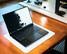 Un ordinateur portable de jeu Razer Blade 16 noir mat, ouvert, posé sur un tapis de bureau blanc posé à son tour sur une table en bois aux teintes chaudes, affichant un fond d'écran en noir et blanc aux couleurs vives.