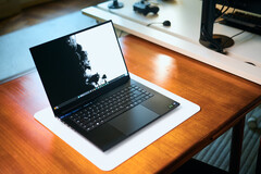 Un ordinateur portable de jeu Razer Blade 16 noir mat, ouvert, posé sur un tapis de bureau blanc posé à son tour sur une table en bois aux teintes chaudes, affichant un fond d'écran en noir et blanc aux couleurs vives.