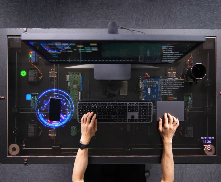 Le bureau « à monter soi-même » de HTX Studio, inspiré de l'AirPower, en situation d'utilisation, avec un téléphone en charge sur l'écran transparent, à côté d'un clavier, d'un pavé tactile et d'un écran.