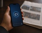 Une main tient un smartphone sur lequel apparaît l'icône d'un chatbot ; un journal imprimé est posé à côté.