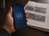 Une main tient un smartphone sur lequel apparaît l'icône d'un chatbot ; un journal imprimé est posé à côté.