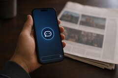 Une main tient un smartphone sur lequel apparaît l'icône d'un chatbot ; un journal imprimé est posé à côté.