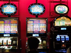 Machines à sous à l'intérieur d'un casino avec des écrans lumineux et des personnes jouant devant (Image source : djedj via Pixabay)