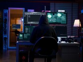 Personnage encapuchonné devant une installation multi-écrans menant une cyberattaque. 