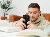 Un adolescent en sweat à capuche tapant sur son smartphone, des manuels scolaires ouverts devant lui