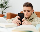 Un adolescent en sweat à capuche tapant sur son smartphone, des manuels scolaires ouverts devant lui