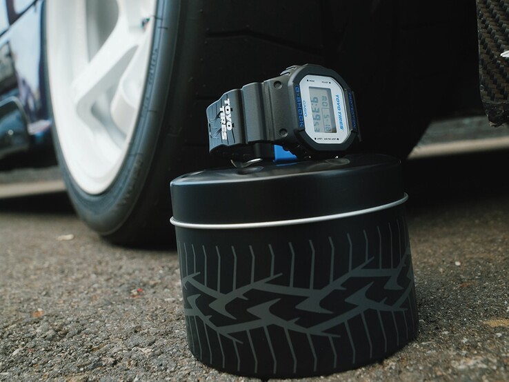 Montre Casio G-Shock DW5600TT25-1 x Toyo Tires présentée sur une boîte métallique sur le thème des pneus, à côté d'une roue de voiture.