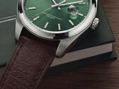 Gros plan de la montre Timex Legacy Day and Date, montrant son cadran vert soleillé, sa lunette argentée et ses cornes brossées, sa couronne striée et son bracelet en cuir marron texturé, sur fond de boîte vert foncé et de surface en bois.