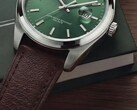 Gros plan de la montre Timex Legacy Day and Date, montrant son cadran vert soleillé, sa lunette argentée et ses cornes brossées, sa couronne striée et son bracelet en cuir marron texturé, sur fond de boîte vert foncé et de surface en bois.