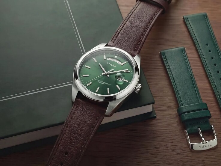 La Timex Legacy Day and Date avec son cadran vert et son bracelet en cuir marron, présentée sur un livre vert foncé à côté du bracelet interchangeable en cuir vert.