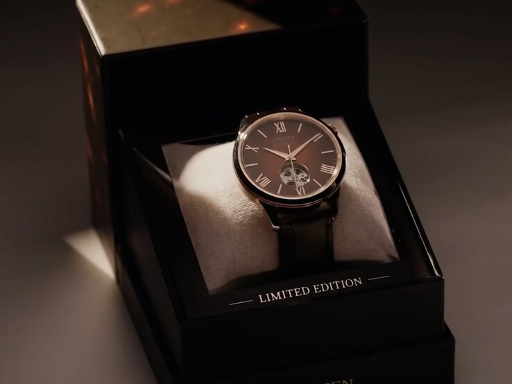 Un plan plus large, éclairé de manière spectaculaire, montrant la montre Citizen posée sur un coussin à l’intérieur de son coffret de présentation sombre à plusieurs niveaux. Un projecteur aux lumières chaudes éclaire le cadran de la montre, et le mot « CITIZEN » est imprimé sur le bord inférieur avant du coffret.