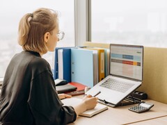 Une femme portant des lunettes travaille sur une feuille de calcul sur son ordinateur portable, avec des classeurs et une calculatrice sur son bureau