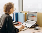 Une femme portant des lunettes travaille sur une feuille de calcul sur son ordinateur portable, avec des classeurs et une calculatrice sur son bureau