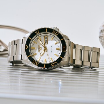 Montre Seiko 5 Sports SKX HDB004 avec un cadran blanc argenté, une lunette tournante noire avec des repères dorés, des aiguilles et des index dorés, et un bracelet en acier inoxydable, photographiée de biais.