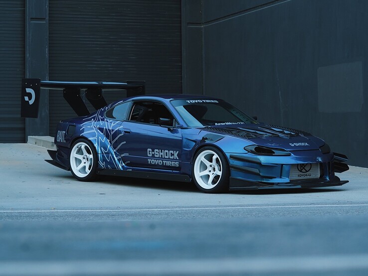 Voiture de drift bleue arborant les graphismes G-Shock et Toyo Tires, garée à l'extérieur d'un bâtiment industriel.