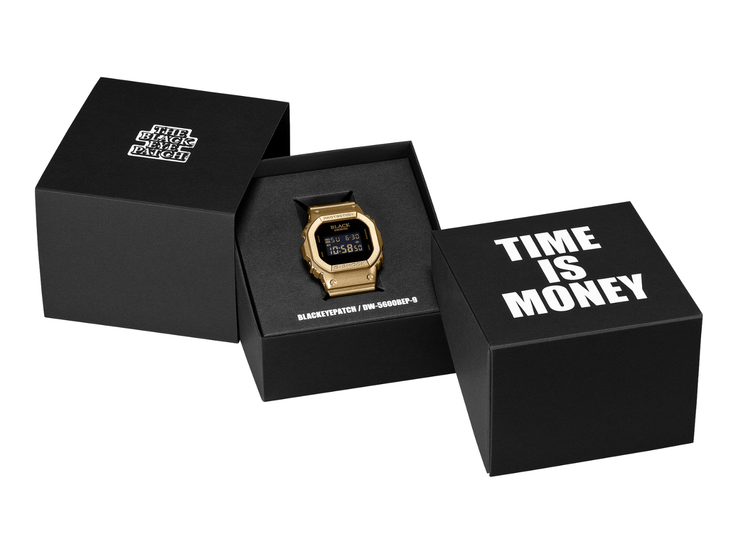 Une montre Casio G-Shock DW-5600BEP-9 dorée, dotée d’un écran numérique noir, est posée entre deux coffrets de présentation noirs. La montre arbore le logo BlackEyePatch sur le cadran, tandis que le coffret au premier plan porte l’inscription « TIME IS MONEY ».