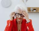 Une femme d’un certain âge, aux cheveux blancs et portant des lunettes, est au téléphone dans un salon lumineux, la main posée sur la tempe.