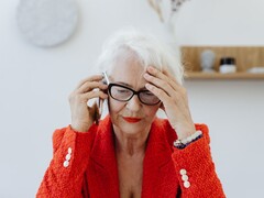 Une femme d’un certain âge, aux cheveux blancs et portant des lunettes, est au téléphone dans un salon lumineux, la main posée sur la tempe.