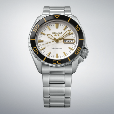 Seiko 5 Sports SKX HDB004 avec un cadran blanc argenté, une lunette noire avec des marquages dorés, des aiguilles et des index dorés, et un bracelet en acier inoxydable, présenté sur un fond gris clair.